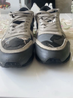Grå sneakers med kamouflagemönster - Säljer ett par sneakers i olika nyanser av grått med svart och vitt kamouflagemönster. Skorna har mockadetaljer, textilpartier och svarta sulor. Klassisk låg modell med snörning och rund tå.