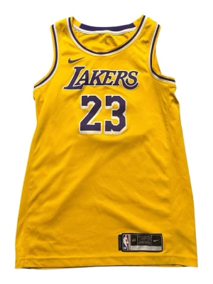 Lakers 23 - Säljer en gul basketlinne med Lakers-tryck och nummer 23 på både fram- och baksidan. På ryggen står det 'JAMES'. Linnet har blå och vita detaljer runt hals och ärmhål. Perfekt för basket eller streetwear.