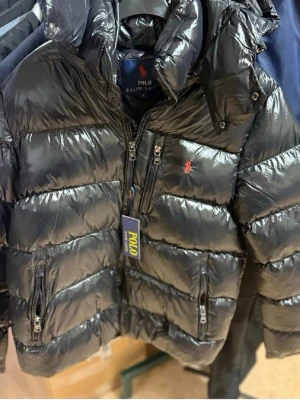 Svart dunjacka från Polo Ralph Lauren - Snygg svart dunjacka från Polo Ralph Lauren med glansig finish och klassisk röd logga på bröstet. Jackan har dragkedja framtill, två sidofickor med dragkedja och en hög krage med huva. Perfekt för kalla vinterdagar.