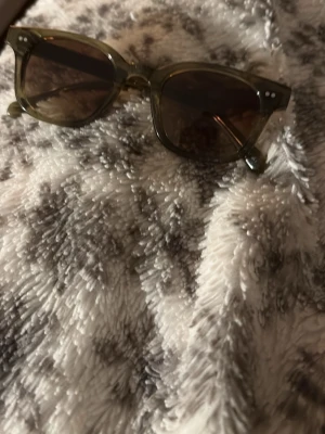 Olivgröna solglasögon i retrostil - Snygga solglasögon med olivgröna, transparenta bågar och mörka glas. Modellen har en klassisk retroform med markerade hörn och små silvriga detaljer vid fronten. Perfekta för dig som vill ha en cool och tidlös look.