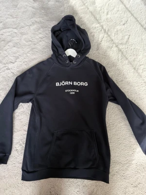 Svart hoodie från Björn Borg - Snygg svart hoodie från Björn Borg med vit text på bröstet där det står 'BJÖRN BORG STOCKHOLM 1956'. Hoodien har en stor ficka framtill och klassisk huva med dragsko. Perfekt för en chill och sportig look.