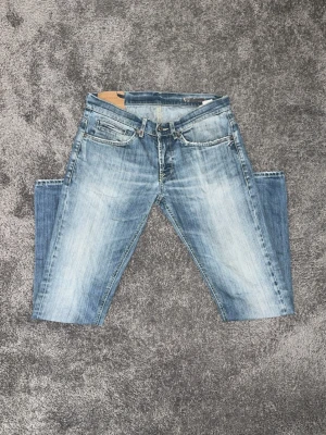 Blå slitna jeans från Dondup - Säljer ett par blå jeans från Dondup med snyggt slitna detaljer och ljus tvätt. Klassisk femficksmodell med Dondup loggan på bakfickorn och Slim-fit passform. Perfekta för en modern och tidlös stil. 