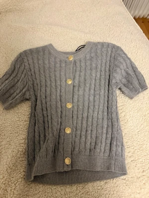 Grå kabelstickad kofta H&M kort ärm - Grå kabelstickad kofta från H&M med korta ärmar och rund hals. Koftan har sex guldfärgade knappar framtill och ribbade muddar vid ärmslut och nederkant. Perfekt för lager på lager och enkel att styla till olika outfits.
