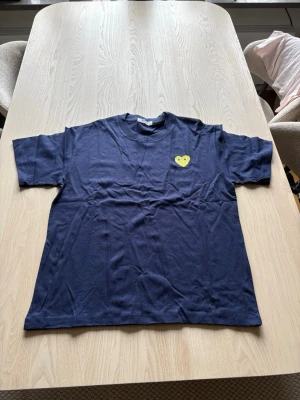 Comme des garcons - Säljer en mörkblå t-shirt från comme des garcons med ett gult hjärta med ögon tryckt på bröstet. T-shirten har rund hals och korta ärmar. Tillverkad i mjuk bomull och har en avslappnad passform. Perfekt för dig som gillar enkel men unik stil.