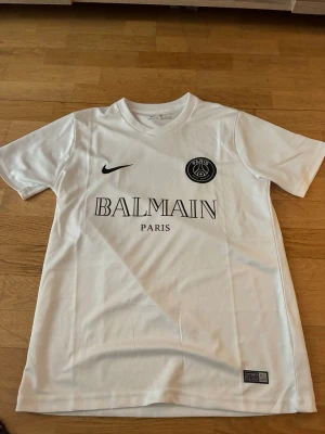 Vit PSG x Balmain t-shirt Nike S - Snygg vit t-shirt från Nike i samarbete med PSG och Balmain. Tröjan har korta ärmar, rund hals och tryck med Balmain Paris och PSG-logga på bröstet. Tillverkad i Dri-FIT-material som andas, perfekt för träning eller sport.