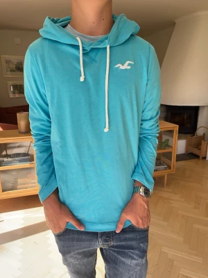 Hollister tunn hoodie herr vintage blå  - Hollister hoodie i snygg turkos/blå färg med diskret vit Hollister-måsen på bröstet. Enkel och stilren design med huva och vita snören. Perfekt för en klassisk Hollister/preppy look.  #Hollister #hoodie #preppy #Y2K #surfstyle #streetwear #college #vintage
