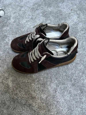 Bruna maison Margiela  - Säljer ett par snygga sneakers i mörkbrun mocka med svarta skinnpaneler och beige snörning. Skorna har en klassisk rund tå och gummisula som ger bra grepp. Perfekta för dig som gillar stilrena och tidlösa skor. Storlek 44. De har två hål i skon.