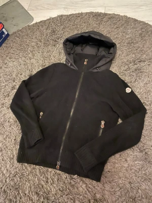 Svart stickad hoodie med dragkedja - Svart stickad hoodie med hel dragkedja och två fickor med dragkedja. Jackan har en vadderad huva och ribbade muddar vid ärmslut och nederkant. Perfekt för kyliga dagar och enkel att matcha med olika outfits.