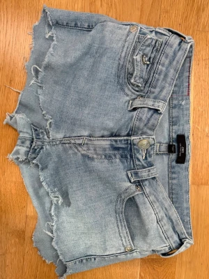 Ljusblå jeansshorts low waist True Religion - Säljer ett par ljusblå jeansshorts från True Religion low waist, fransiga kanter och klassiska fickor både fram och bak. Shortsen har synliga sömmar, knappgylf och detaljerade bakfickor med lock och metallknappar. Perfekta för sommaren!