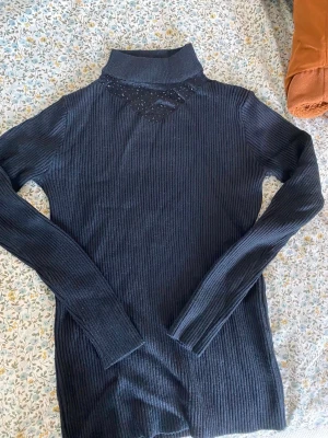 Svart ribbstickad polotröja - Snygg svart ribbstickad polotröja med lång ärm och hög krage. Tröjan har ett diskret meshparti med små glittriga detaljer vid halsen som ger en cool touch. Perfekt för lager på lager och passar till allt. Materialet känns mjukt och stretchigt.