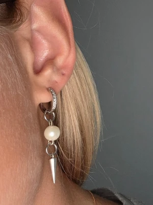 Silvriga örhängen med pärlor och spikes - Säljer ett par trendiga silvriga örhängen med vita pärlor och spetsiga spikes. Örhängena har små gnistrande stenar på ringen och en cool kombination av klassisk pärla och edgy metallspets. Perfekta för dig som vill sticka ut med din stil.