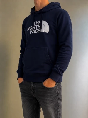Mörkblå hoodie från The North Face - Snygg mörkblå hoodie från The North Face med stor vite logga på bröstet. Hoodien har huva med dragsko och en stor magficka framtill. Tillverkad i mjukt material och har ribbade muddar vid ärmslut och nederkant. Perfekt för en avslappnad och trendig stil.