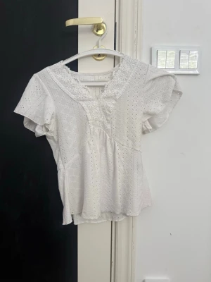 Vit broderad blus med spetsdetaljer - Söt vit blus med broderade hålmönster och spetsdetaljer vid halsringningen. Blusen har en lätt v-ringning, korta ärmar och en avslappnad passform med volang nedtill. Perfekt för en somrig och romantisk look.