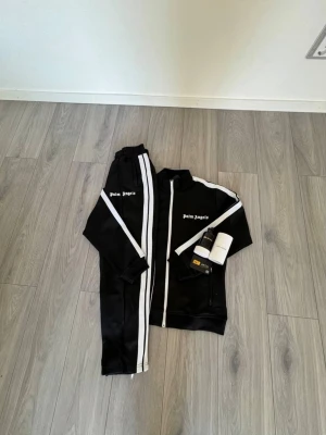 Svart tracksuit från Palm Angels - Säljer en svart tracksuit från Palm Angels med vita ränder längs ärmar och ben. Setet består av zip-jacka och byxor, båda med logga på bröst och lår. Materialet är mjukt och stretchigt, perfekt för chill eller loungewear. Klassisk sportig look med ribbade muddar.