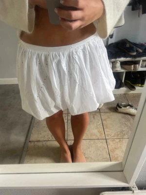 Vit kort kjol med volang - Söt vit kort kjol med volang och broderade detaljer. Kjolen har en luftig och puffig form med resår i midjan, vilket ger en bekväm passform. Perfekt för varma dagar och somriga outfits.