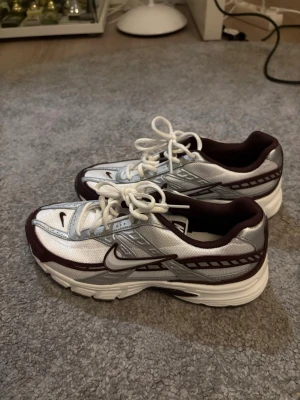 Nike silver och vinröda sneakers - Snygga sneakers från Nike i silver och vinrött med vita detaljer. Skorna har en sportig design med mesh och syntetmaterial, vita snören och en rejäl sula för extra komfort. Perfekta för dig som gillar retrovibbar och streetstyle. Oanvända