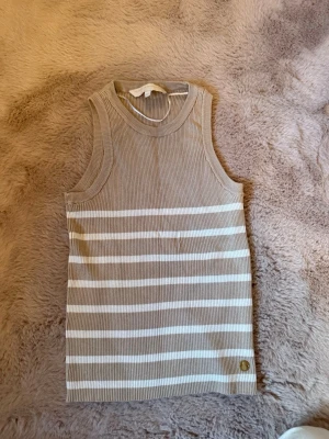 Beige och vit randig ribbad linnetopp - Säljer en ribbad linnetopp i beige och vitt med horisontella  ränder. Toppen är ärmlös och har en rund halsringning. Skönt och stretchigt material 🤍