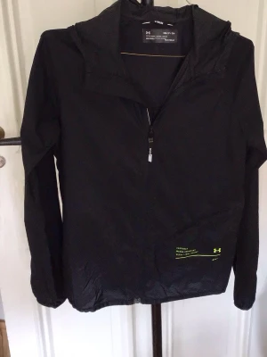 Svart löparjacka från Under Armour - Snygg svart löparjacka från Under Armour med halv dragkedja framtill och huva. Tillverkad i lätt och funktionellt HeatGear-material som andas. Diskreta loggor och detaljer i neon på framsidan. Perfekt för löpning och träning utomhus.
