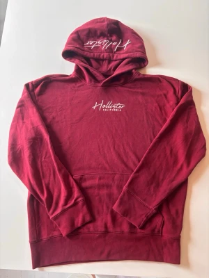 Vinröd hoodie från Hollister - Säljer en vinröd hoodie från Hollister med vit broderad logga på bröstet och huvan. Perfekt för en av kopplad stil som passar till till alla stilar. Storlek L, 