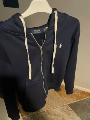 Marinblå hoodie från Polo Ralph Lauren - Snygg marinblå hoodie från Polo Ralph Lauren med dragkedja framtill, vit broderad logga på bröstet och vita snören i huvan. Hoodien har fickor framtill och är tillverkad i mjukt material som känns skönt mot huden. Perfekt för en avslappnad och stilren look.