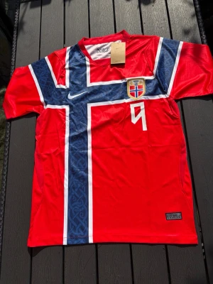 Norge fotbollströja Haaland #9 - Röd Norge fotbollströja från Nike med blått och vitt kors, landslagets emblem och nummer 9 på framsidan. På ryggen står det 'Braut Haaland' och stort nummer 9. Tillverkad i lätt och ventilerande material, perfekt för match eller träning.