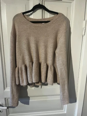 Beige volangstickad tröja Gina Tricot - Supersöt beige stickad tröja från Gina Tricot med volangdetalj nedtill. Tröjan har rund halsringning, långa ärmar och ribbstickade muddar. Perfekt för dig som gillar en feminin och trendig look. Materialet känns mjukt och skönt mot huden.