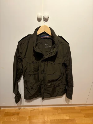 Olivgrön vindjacka från Snoot - Snygg olivgrön vindjacka från Snoot med hög krage, flera fickor framtill och axelklaffar. Jackan har dragkedja och knappar samt är tillverkad i ett lätt, vindtätt material. Perfekt för dig som vill ha en stilren och funktionell jacka.