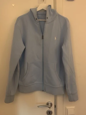 Ljusblå hoodie från Polo Ralph Lauren - Snygg ljusblå hoodie med dragkedja från Polo Ralph Lauren. Klassisk design med huva, fickor framtill och den ikoniska broderade loggan på bröstet. Perfekt för en chill och stilren look. Mjuk och skön bomullsmix som passar året runt.