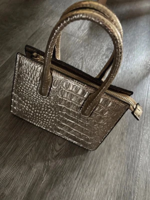 Guldig handväska i krokopräglat material - Snygg handväska i metallic guld med krokopräglat mönster. Från H&M. Väskan har två stadiga handtag och dragkedja upptill. Stilren och fyrkantig form som ger en trendig look. Perfekt accessoar för att lyfta din outfit. Oanvänd men finns ingen prislapp kvar! 