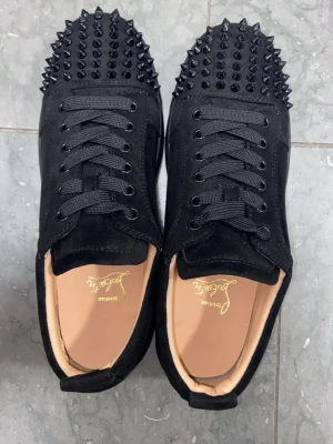 Svarta Loubs Med Spikar - Säljer ett par svarta sneakers från Christian Louboutin med mattsvarta nitar över tån och klassisk röd sula. Skorna har svarta skosnören, ovandel i mocka och en låg silhuett. Perfekta för dig som vill sticka ut med en edgy och lyxig stil.