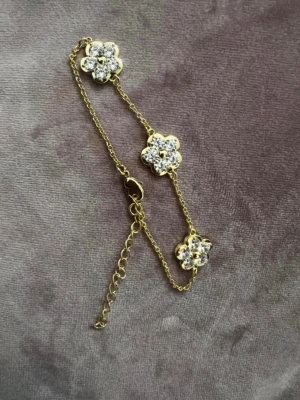 Guldarmband med blommor och pärlor - Snyggt armband i guldton med tre blommor dekorerade med gnistrande pärlor. Kedjan är tunn och har ett justerbart lås för perfekt passform. Blommorna ger en söt och trendig touch till din stil.