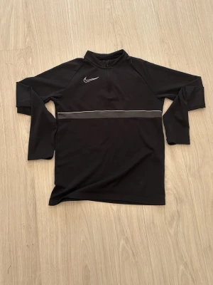 Nike långärmad träningströja  - Svart långärmad träningströja från Nike med halv dragkedja framtill och vit broderad logga på bröstet. Tröjan har en diskret vit linje över bröstet och är tillverkad i ett mjukt, stretchigt funktionsmaterial som passar perfekt till träning.