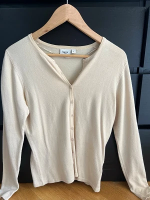 Beige kofta från Saint Tropez - Snygg beige kofta från Saint Tropez med v-ringning och knäppning framtill. Långärmad och i mjukt, tunt material som passar perfekt till lager på lager. Enkel och stilren design som är lätt att matcha med olika outfits.
