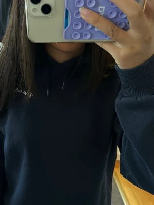 Mörkblå hoodie med huva - Säljer en mörkblå hoodie med huva och snören. Snörena är ganska korta. Hoodien har ribbade muddar vid ärmsluten och ett tryck i vitt på bröstet. Perfekt för dig som gillar en enkel och stilren look.