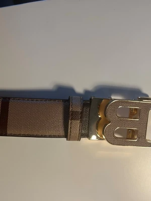 Beige och brunt randigt BB bälte - Snyggt bälte i beige och brunt med randigt mönster och en stor metallspänne i guld och silver. Bältet har en bred design och passar perfekt för att lyfta vilken outfit som helst.