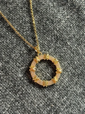 Guldigt halsband med cirkelhänge - Snyggt halsband i guldfärg med ett runt hänge dekorerat med små gnistrande stenar och detaljer som påminner om taggtråd. Kedjan är tunn och elegant, och hänget har en unik design som verkligen sticker ut. Perfekt accessoar för dig som gillar statement-smycken.