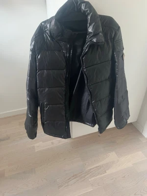 Svart pufferjacka med emblem - Snygg svart pufferjacka med hög krage och quiltad design. Jackan har dragkedja framtill och en ficka med lock på ärmen, dekorerad med ett runt tygmärke där det står 'Fashion W'. Materialet är glansigt och ger en modern look.