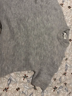Grå stickad tröja Gina Tricot - Mysig grå stickad tröja från Gina Tricot i ullblandning. Tröjan har korta ärmar, rund halsringning och ribbstickade detaljer vid ärmslut och nederkant. Perfekt för lager-på-lager och enkel att matcha med olika outfits.