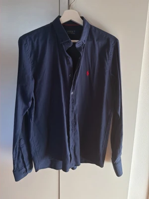 Mörkblå skjorta från Polo Ralph Lauren - Snygg mörkblå skjorta från Polo Ralph Lauren med klassisk button-down krage och röd broderad logga på bröstet. Skjortan har långa ärmar och knäppning framtill. Tillverkad i mjukt bomullstyg som känns bekvämt hela dagen. Köptes i Kroatien vet ej om äktheten