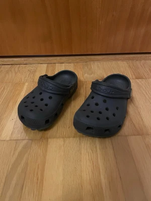 Svarta Crocs tofflor med rem - Svarta Crocs tofflor i klassisk modell med justerbar hälrem och ventilerande hål på ovansidan. Tillverkade i lätt och bekvämt gummimaterial som är enkelt att rengöra. Perfekta för chill dagar hemma eller när du vill ha något snabbt att slänga på fötterna.