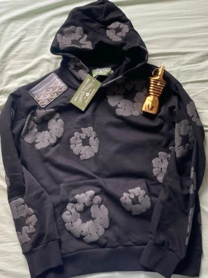 Svart hoodie från Denim Tears - Säljer en svart hoodie från Denim Tears med gråa blommönster över hela tröjan. Hoodien har en klassisk huva och magficka framtill. Materialet är mjukt och bekvämt, perfekt för en avslappnad streetwear-look.