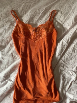 Ribbad orange body med spets - Snygg orange body med tunna axelband och ribbad struktur. Överdelen har spetsdetaljer och en liten rosett framtill. Materialet ser mjukt och stretchigt ut, perfekt för lager på lager eller som statement-plagg.