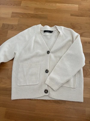 Vit kofta från Vero Moda - Mysig vit kofta från Vero Moda med stora knappar framtill och två rejäla fickor. Koftan har ribbade muddar vid ärmslut och nederkant samt en avslappnad passform. Perfekt att slänga på sig när det är lite kyligt ute.