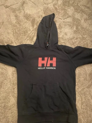 Svart hoodie från Helly Hansen - Säljer en svart hoodie från Helly Hansen med stor röd HH-logga och vit text på bröstet. Hoodien har en klassisk känguruficka framtill och huva. Perfekt för dig som gillar streetwear och vill ha en enkel men snygg look. En som vill köpa nu så skynda er ifall ni vill köpa