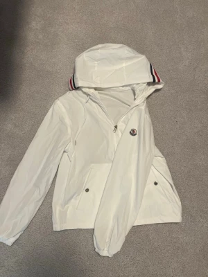 Vit vindjacka från Moncler - Snygg vit vindjacka från Moncler med dragkedja framtill, huva med röd och marinblå randdetalj samt två fickor med tryckknappar. Jackan har ett broderat Moncler-märke på bröstet och är tillverkad i ett lätt, vattenavvisande material.