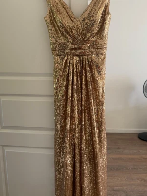 Lång glittrig guldig klänning - Säljer en elegant långklänning i guld med paljetter över hela klänningen. Klänningen har v-ringning både fram och bak samt markerad midja för en snygg siluett. Perfekt för dig som vill sticka ut på bal eller fest.