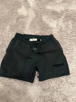 Svarta shorts Essentials Fear of God - Säljer ett par svarta shorts från Essentials Fear of God med snörning i midjan och logga på vänster ben. Shortsen har en avslappnad passform och är perfekta för sommaren. Tillverkade i mjuk bomull för maximal komfort.