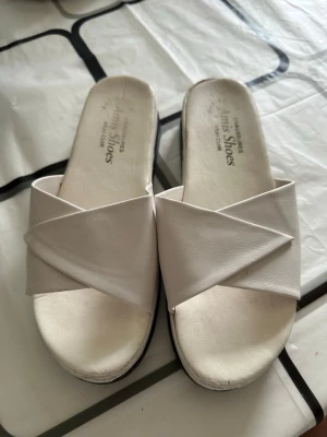 Vita sandaler från Amis Shoes - Säljer ett par vita sandaler från Amis Shoes i äkta läder. De har en öppen tå och breda korslagda remmar över foten. Snygg och enkel design som passar till många olika outfits. Sulan är platt och bekväm.