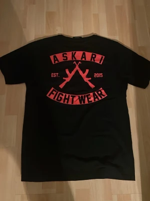 Svart Askari Fight Wear t-shirt - Säljer en svart t-shirt från Askari Fight Wear med rött tryck både fram och bak. Stora korsade gevär och texten 'Askari Fight Wear' ger en tuff look. Klassisk passform och rund hals. Perfekt för dig som gillar streetwear.