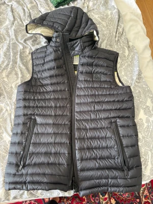 Moose Knuckle väst. Jätte bra skick, knappt använd.  - Snygg svart dunväst med avtagbar huva och dragkedja framtill. Västen har två framfickor med dragkedja och quiltad design. Insidan är ljus med kontrastfärg. Perfekt för lager-på-lager och håller dig varm under kyliga dagar.